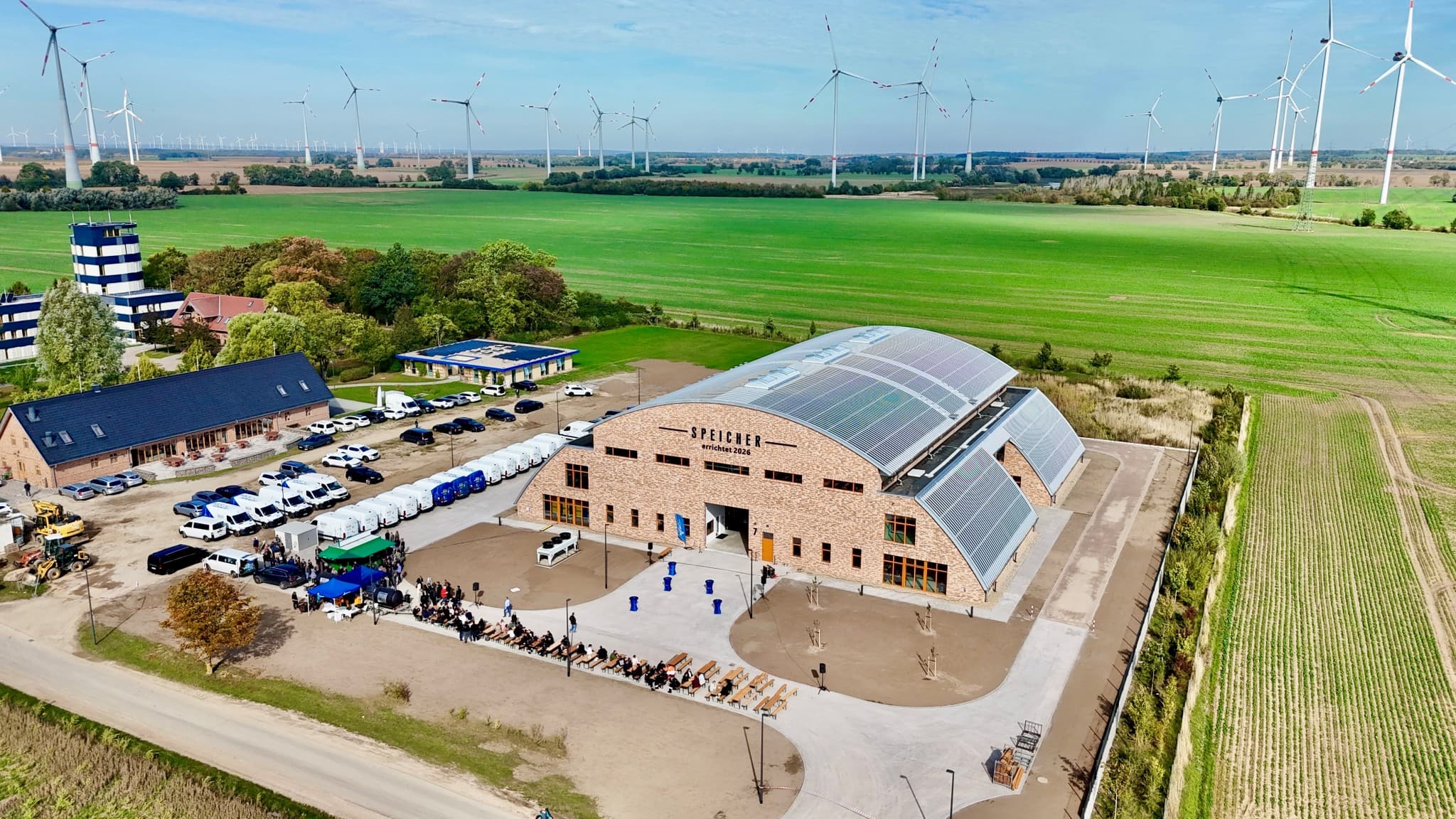 Luftaufnahme des neuen ENERTRAG Speichers am Energiecampus Dauerthal, umgeben von grünen Feldern und Windkraftanlagen, mit Besuchern zur Eröffnung vor dem Gebäude.