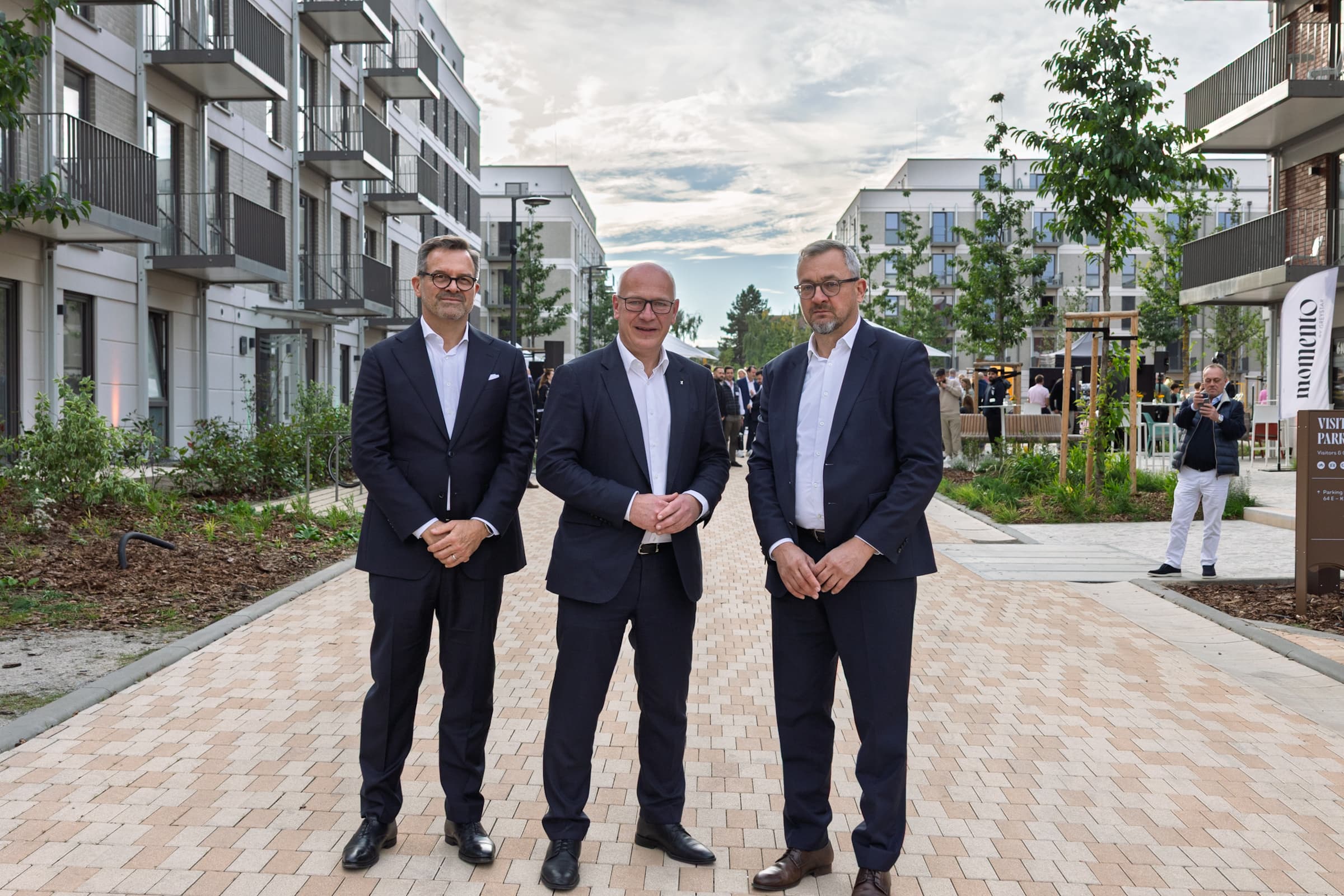 Drei Männer in Anzügen stehen bei der Eröffnung von Momento Berlin im neuen Wohnquartier in Berlin-Neukölln zusammen.