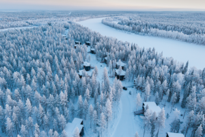 Luftaufnahme von Noa Villas im verschneiten Lappland, Finnland, mit mehreren Winterhütten zwischen schneebedeckten Bäumen und einem zugefrorenen Fluss.
