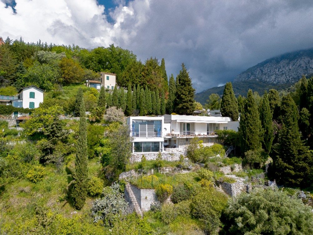 Außenansicht der Villa Tempe à Pailla in Menton, ein modernistisches Gebäude, umgeben von Bäumen und Bergen.